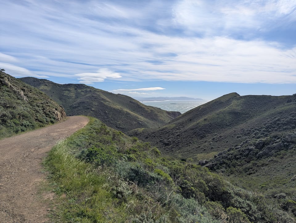 a photo shows a bay area landscape with a wide dirt hiking path and rolling green hills. There is a glimpse of ocean past the hills, and a blue sky streaked with white clouds